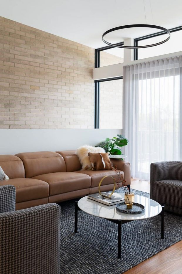 Bright contemporary living room with a caramel leather sectional, patterned armchairs, a round marble-top coffee table, and a dark textured area rug. A pale exposed brick feature wall contrasts with large black-framed windows and sheer curtains, while a simple circular pendant light hangs above the seating area.