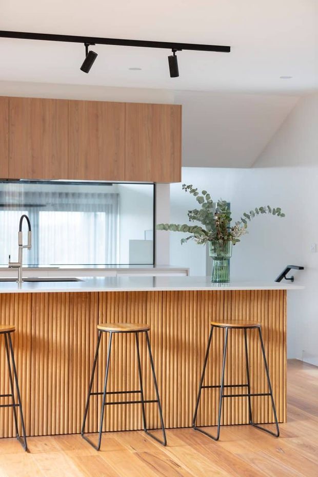 Bright contemporary kitchen featuring a large island with a vertically fluted timber-clad front, white benchtop, integrated sink and tapware, tall timber cabinetry, and black metal bar stools. A large mirror reflects a window and sheer curtains, while black track lighting and warm timber flooring add contrast.