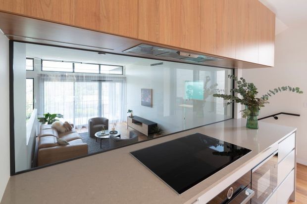 Contemporary open-plan kitchen viewed from the island, featuring warm timber overhead cabinetry, a light stone benchtop, black induction cooktop, and white lower cabinetry. A large glazed partition opens the kitchen visually to a bright living area with a sofa, coffee table, television console, and indoor greenery.