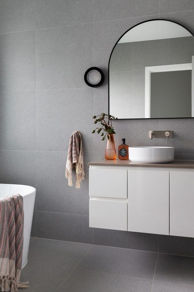 A contemporary bathroom featuring large-format grey tiled walls and floor, a floating white vanity with a taupe stone-look benchtop, a white vessel basin, wall-mounted tapware, and a large arched black-framed mirror. A freestanding white bathtub is partially visible at left, with a towel and decorative accessories adding warmth.