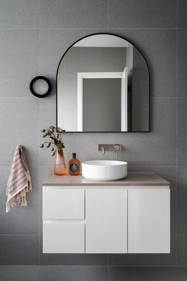 A compact contemporary bathroom vanity featuring a white wall-mounted cabinet with a taupe-grey benchtop, white vessel basin, wall-mounted tapware, and a large arched black-framed mirror. The walls and floor are clad in large-format grey tiles, with a round black wall light, towel, and small decorative accessories.