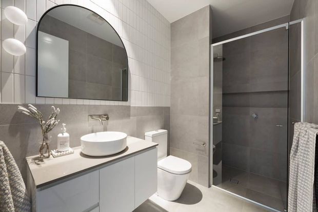 Contemporary bathroom with a wall-mounted vanity, white vessel basin, arched black-framed mirror, concealed wall-mounted tap, toilet, and frameless glass shower enclosure. The room uses large grey tiles with a white small-format tiled upper wall and neutral modern finishes.