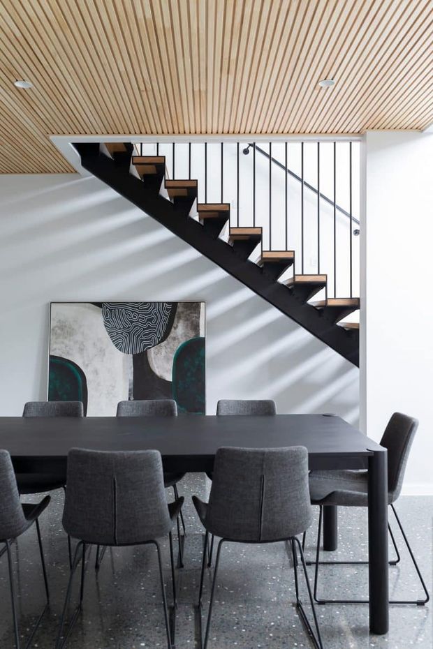 A contemporary dining area beneath a striking open staircase. The room features a large dark dining table with grey upholstered chairs, a black steel stair structure with timber treads, a vertically slatted timber ceiling, white walls, terrazzo flooring, and a monochrome abstract artwork. Slim black vertical balusters and a simple metal handrail create a graphic architectural backdrop.