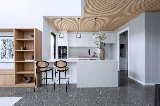 Bright contemporary kitchen with a large pale stone island, white flat-panel cabinetry, integrated appliances, timber slatted ceiling, terrazzo-style flooring, black-framed bar stools, and open timber shelving. A hallway or pantry opening is visible to the right.