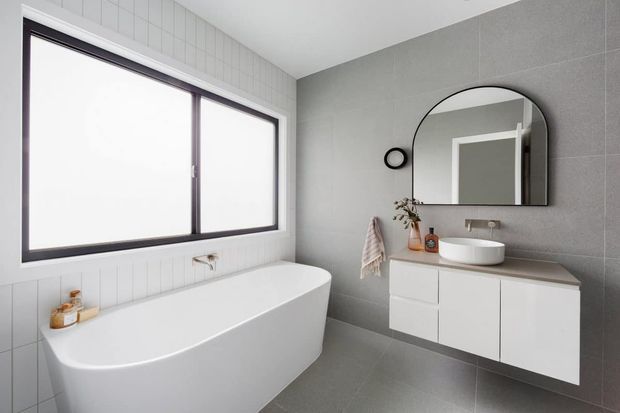 A bright contemporary bathroom featuring a freestanding white bathtub beneath a large dark-framed window, grey tiled walls and floor, a floating white vanity with a grey benchtop, round vessel basin, and a large arched mirror.