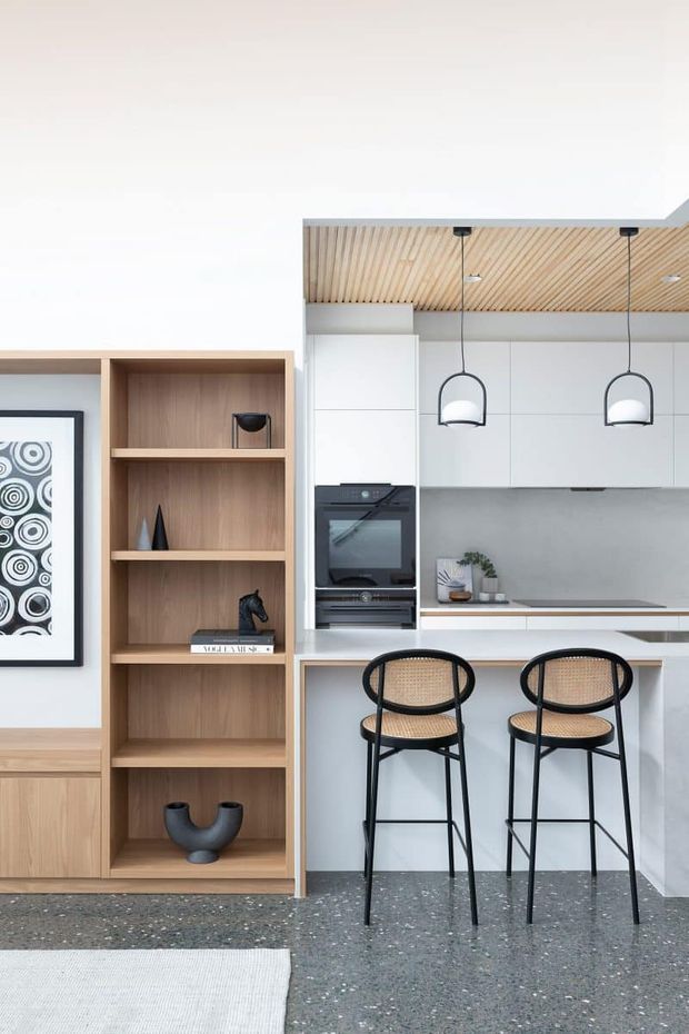 Bright contemporary kitchen and living area with a white flat-panel kitchen, integrated appliances, pale timber shelving and cabinetry, a terrazzo-style floor, and a slim island with two black-framed woven bar stools. The ceiling features linear timber battens, with simple pendant lights above the island. A built-in timber display shelf and framed artwork occupy the adjoining wall.