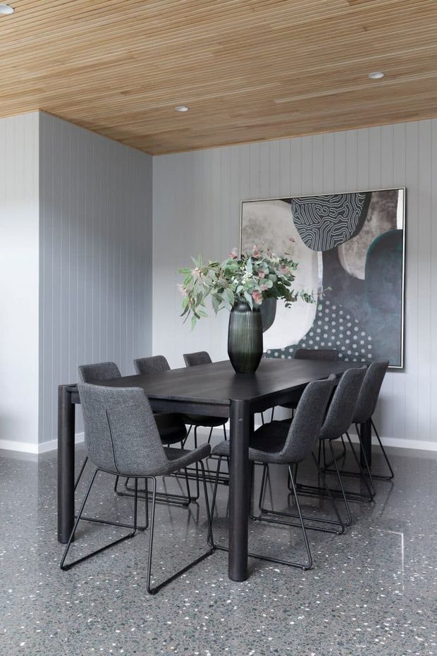 A contemporary dining area with a dark rectangular dining table surrounded by upholstered charcoal dining chairs. The room has vertically panelled light-grey walls, a warm timber slatted ceiling with recessed downlights, and a polished terrazzo floor. A large abstract artwork and a black vase with foliage provide visual accents.