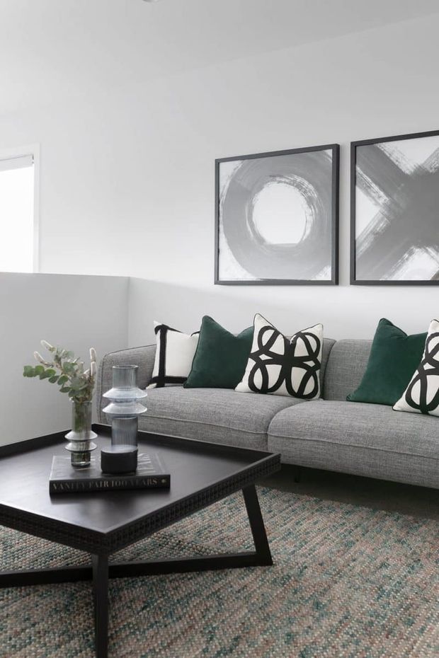 A bright, contemporary living room featuring a large grey upholstered sectional sofa with dark green and monochrome patterned cushions. A black rectangular coffee table sits over a textured patterned rug, with minimalist glass and metal decor. White walls and two large abstract monochrome artworks create a clean, graphic interior.