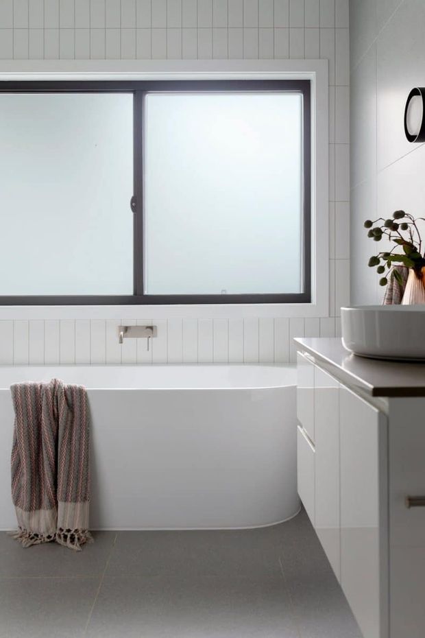 A bright, contemporary bathroom with a freestanding white bathtub beneath a large frosted window. White vertical wall tiles, grey floor tiles, a wall-mounted chrome tap, white vanity cabinetry, a dark benchtop, and a vessel basin are visible. A textured towel and small potted plant add muted accents.