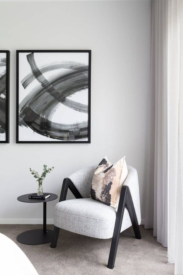 A bright, refined corner of a contemporary living room featuring a light upholstered armchair with dark angled timber arms, a small round black side table, abstract black-and-white artwork in black frames, sheer white curtains, and a neutral carpet. The space has a restrained monochrome palette with soft natural light and minimal styling.