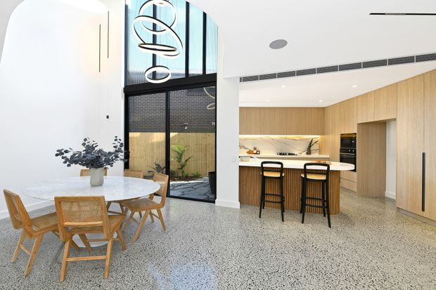 A bright contemporary open-plan dining and kitchen interior with a dramatic double-height void. The space features a round light stone dining table with timber and woven chairs, a polished terrazzo floor, pale timber kitchen cabinetry, a white island benchtop with two bar stools, black-framed glazing, and a sculptural pendant light. A courtyard is visible through the glazing.