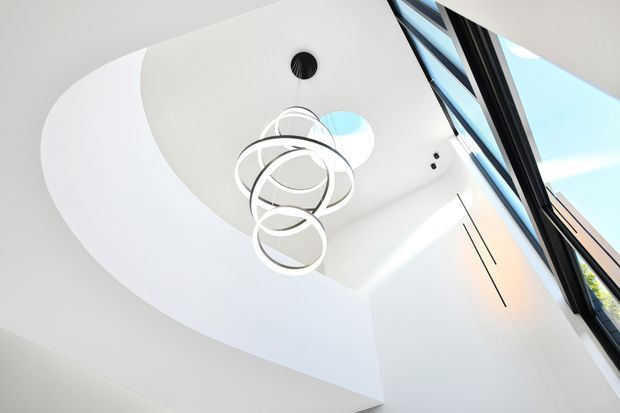 Upward-looking view of a bright contemporary interior with a sculptural circular pendant light suspended beneath a curved white ceiling. Tall black-framed glazing along the right side brings in daylight and emphasizes the high, open volume.