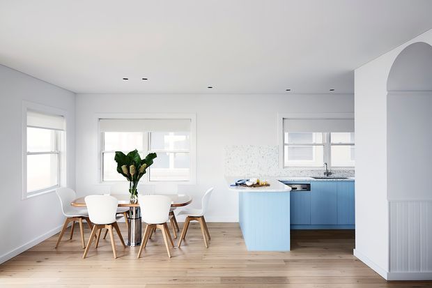 Bright open-plan kitchen and dining area with pale timber flooring, white walls, multiple windows, and a compact light-blue kitchen island. The dining area has a round table with contemporary white chairs, while the kitchen includes blue cabinetry, a white benchtop, stainless-steel appliances, and a tiled splashback. A curved archway and white wall panelling add architectural detail.
