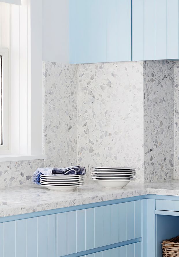Bright, compact kitchen corner with pale blue shaker-style cabinetry, a white and grey terrazzo benchtop and matching terrazzo splashback. A white-framed window provides natural light, while stacked white plates and folded blue-striped textiles sit on the counter. The cabinetry includes vertical fluted detailing and a lower open storage niche.