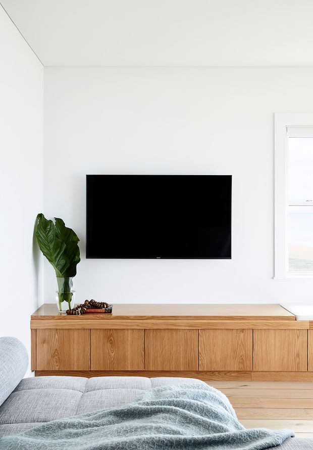 Bright, minimalist living room with a wall-mounted black television above a long, built-in natural timber media console. The room has white walls and ceiling, pale timber flooring, a light grey sofa in the foreground, a soft grey throw, and a tall leafy plant in a glass vase. A white-framed window brings in natural light from the right.