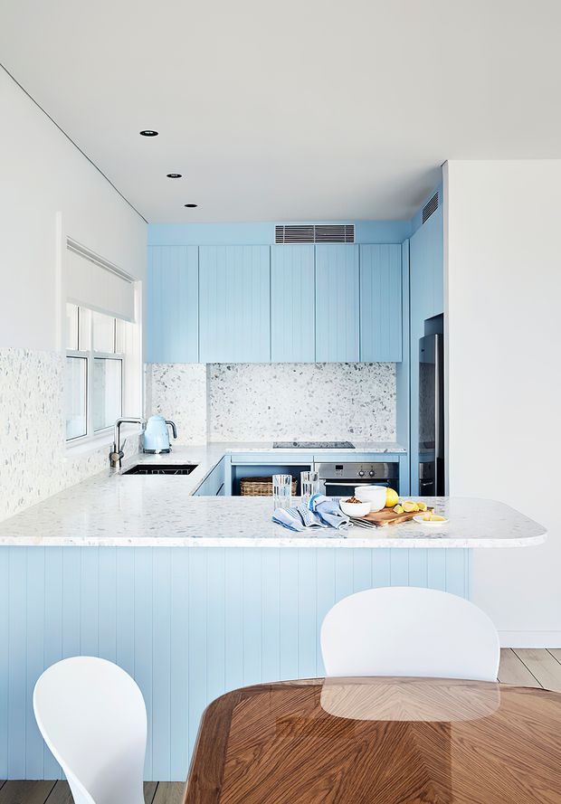 Bright contemporary kitchen with pale blue vertical-panel cabinetry, a white and grey speckled stone benchtop and splashback, integrated appliances, and a peninsula opening toward a dining area. White sculptural dining chairs and a dark timber chevron dining table are visible in the foreground. The space has recessed ceiling lights, light timber flooring, and a clean coastal-inspired palette.