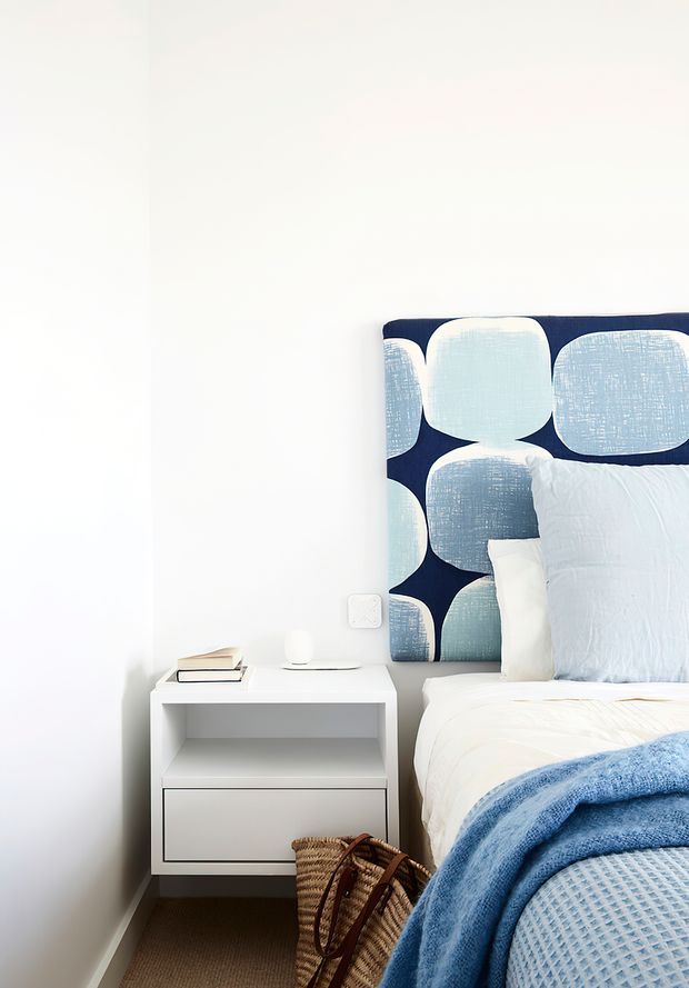 A bright, neatly styled bedroom corner with a white wall, a contemporary built-in white bedside table, and a bed featuring a bold navy and pale blue rounded-pattern upholstered headboard. Pale blue bedding and a textured blue throw create a cool, fresh palette, with books, a small lamp, and a woven basket adding subtle detail.