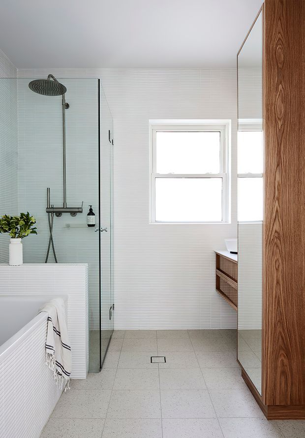 Bright contemporary bathroom featuring a frameless glass walk-in shower with a rain showerhead, freestanding bath partially visible at left, white vertically ribbed wall tiles, pale terrazzo-style floor tiles, a timber vanity and tall timber storage cabinet, and a window providing natural light.