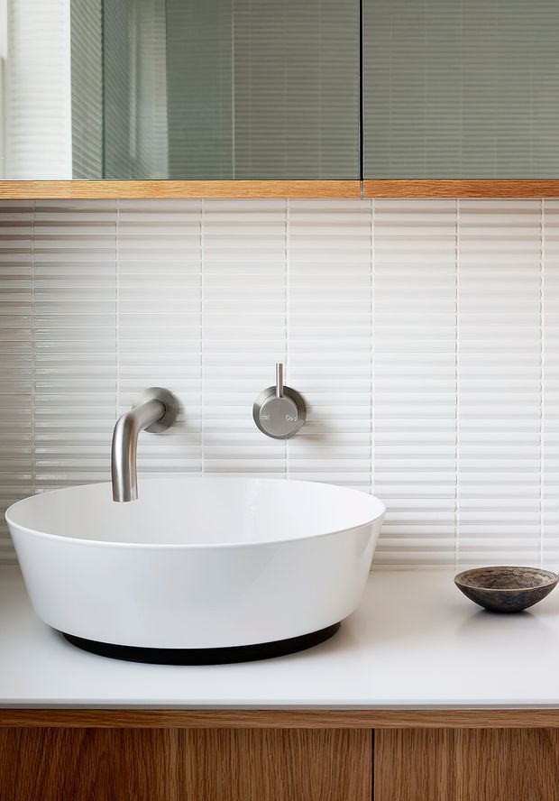 Close-up of a contemporary bathroom vanity featuring a white vessel basin on a white benchtop, brushed metal wall-mounted tapware, slim white horizontal tiled splashback, mirrored cabinetry with timber trim, and a timber vanity base.