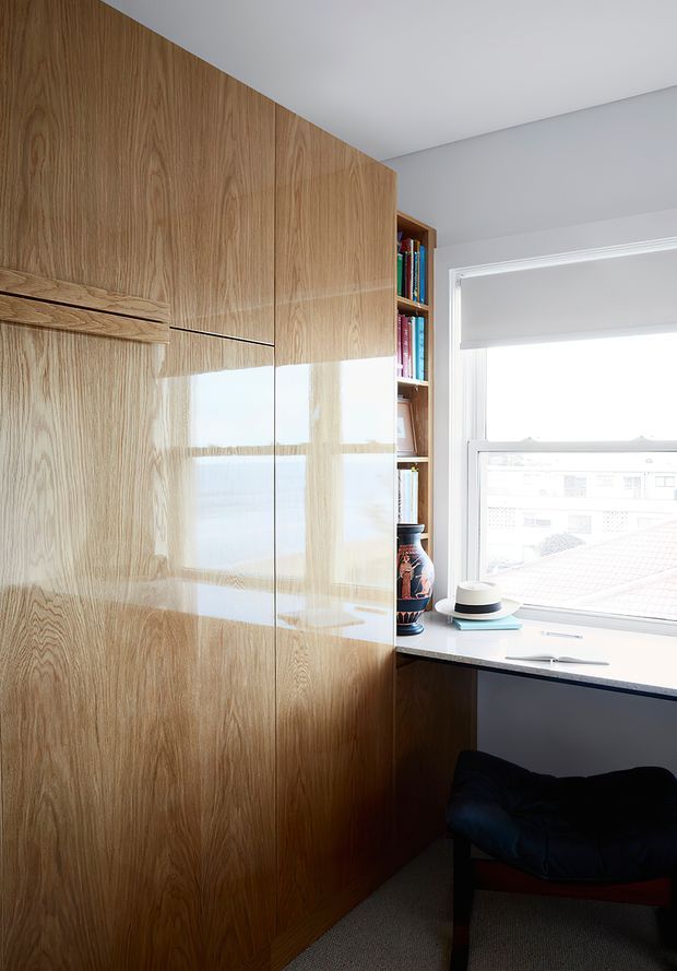 A compact, bright study nook or home office with floor-to-ceiling natural timber built-in cabinetry, integrated shelving, and a white desk beneath a large window. A dark upholstered chair, books, ceramic vessel, and roller blind complete the space. The cabinetry has a warm wood grain and glossy reflective finish, while the walls and ceiling are white.