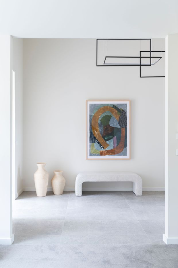 A bright, minimalist entry hall with pale grey walls and large light-grey floor tiles. A sculptural white upholstered bench sits beneath a framed abstract artwork, flanked by two cream ceramic vases. A linear black geometric pendant light hangs from the ceiling, and a carpeted foreground leads into the tiled space.