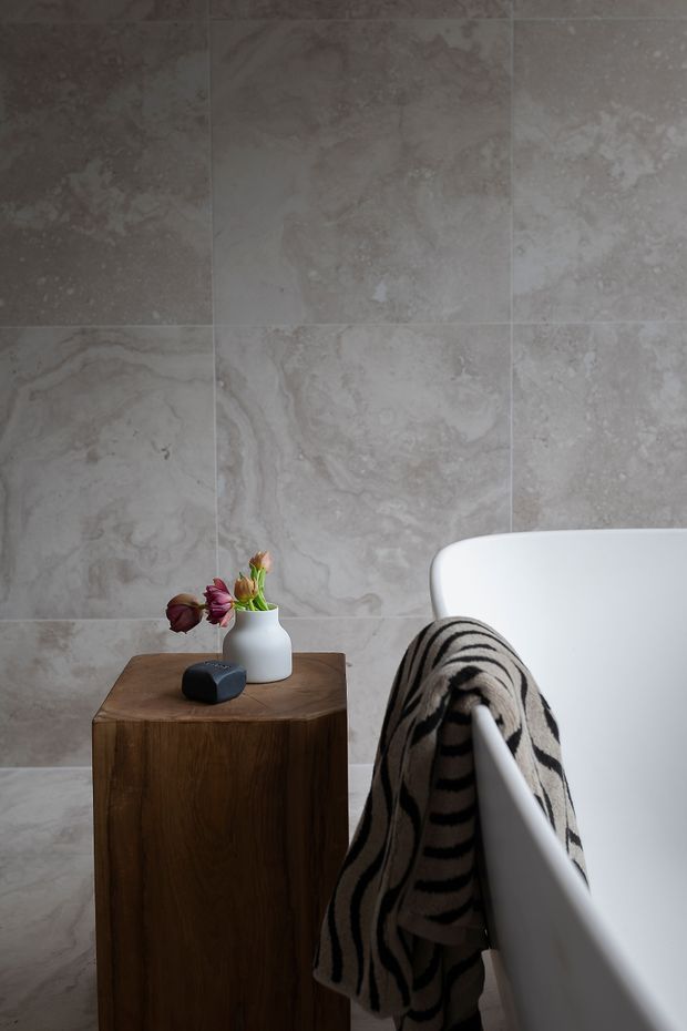 A close-up of a refined bathroom with large-format grey-beige stone-look wall tiles, a white freestanding bathtub, and a dark timber side table. A striped neutral-toned towel hangs over the tub, with a small vase of flowers and soap on the table. The composition is calm and tactile, highlighting the bath setting and material palette.