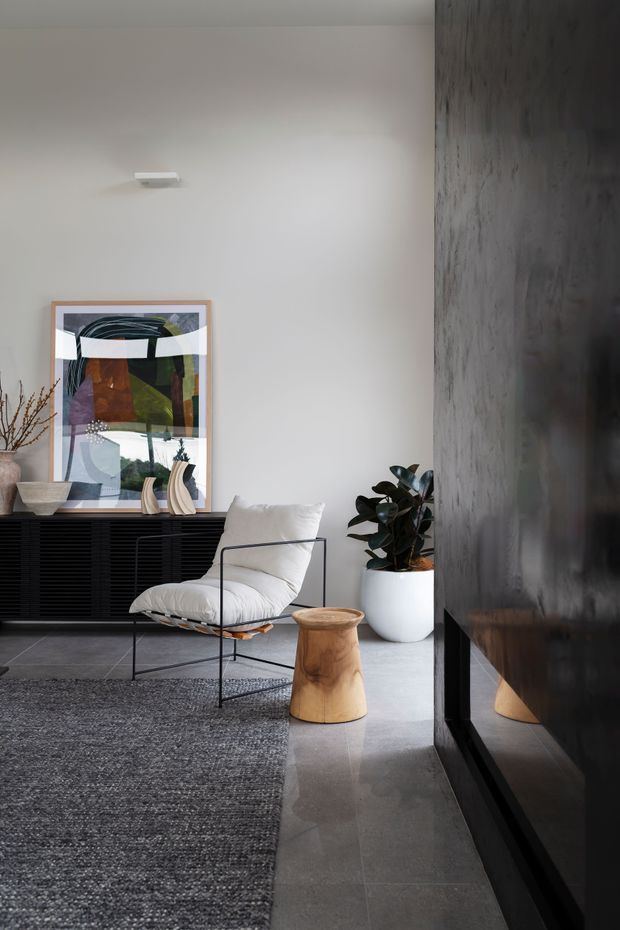 A refined contemporary living room with a neutral palette, polished grey stone flooring, a large textured grey rug, and a sculptural black sideboard. The room features a low white lounge chair, a natural timber stool, a white planter with greenery, abstract framed artwork, and a dark reflective feature wall or joinery element.
