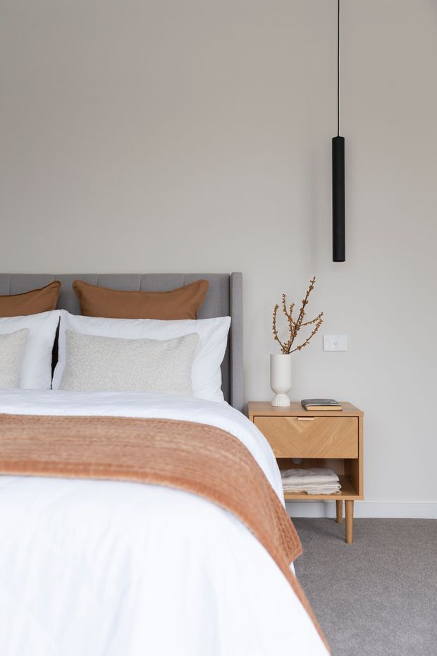 A softly styled modern bedroom with a grey upholstered bedhead, layered white bedding, warm tan cushions and throw, and a timber bedside table with open shelving. A minimal black pendant light hangs beside the bed, complemented by a neutral wall, grey carpet, and simple ceramic vase with dried branches.