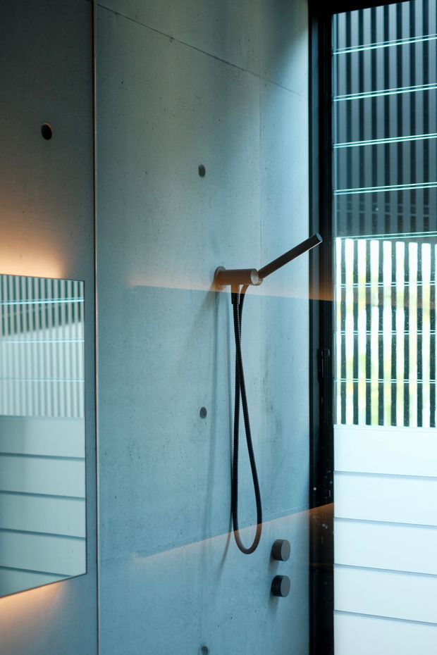 Close-up of a contemporary shower installation with a raw concrete wall, minimalist dark shower hardware, and black-framed glazing with vertical screening. The space has a cool, refined industrial character with natural light entering through the window.