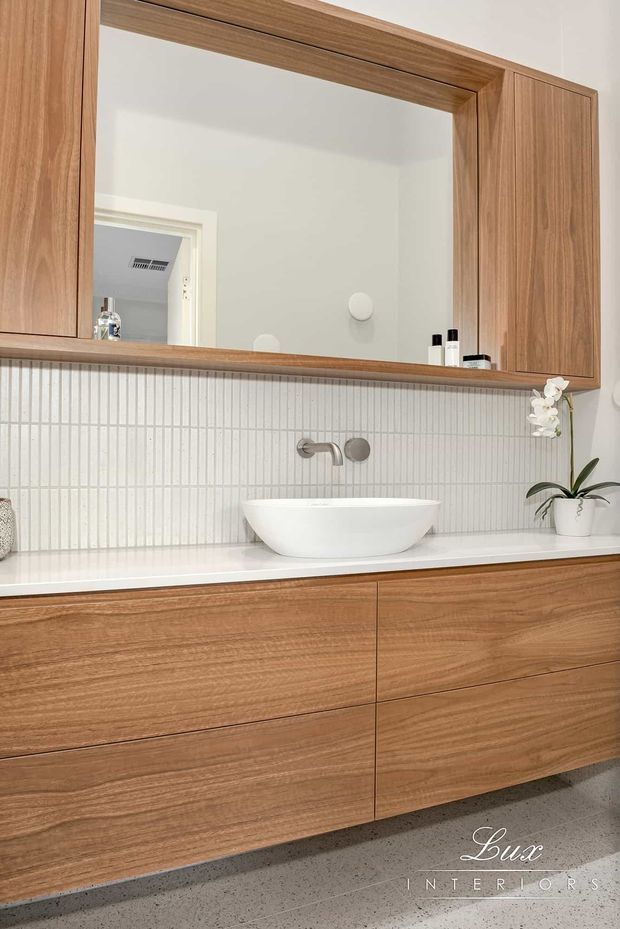 Contemporary bathroom vanity featuring a wall-mounted timber-look cabinet with integrated mirror storage, white engineered-stone benchtop, vessel basin, brushed metal wall-mounted tapware, and a vertically ribbed white tile splashback. The space is bright, clean, and refined, with a small potted orchid adding a natural accent.