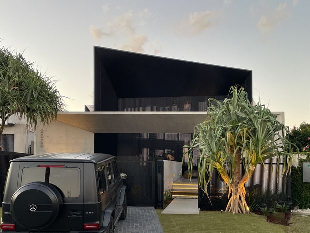 Front exterior view of a contemporary two-storey residence with a dramatic deep black projecting upper facade and broad horizontal concrete-toned canopy. The home has dark vertical metal screening, a gated entry with steps, a paved driveway, lawn, and tropical architectural planting. A vehicle is parked in the foreground.