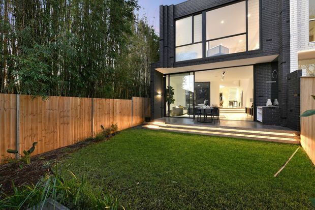 Rear exterior view of a contemporary multi-storey townhouse with a dark brick facade, large black-framed windows, and expansive ground-floor glazing opening onto a stepped patio. A timber privacy fence encloses the landscaped lawn, with dense planting along the boundary. The interior living and dining area is visible through the glass doors.