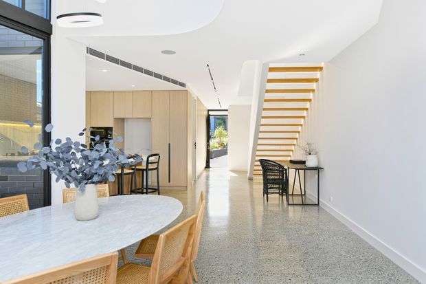 Bright contemporary open-plan interior featuring a dining area in the foreground, a light timber kitchen with island seating, and a hallway leading to a staircase with open timber treads. The space has white curved ceilings and walls, large black-framed glazing, pale timber joinery, polished light terrazzo flooring, and a neutral, airy palette.