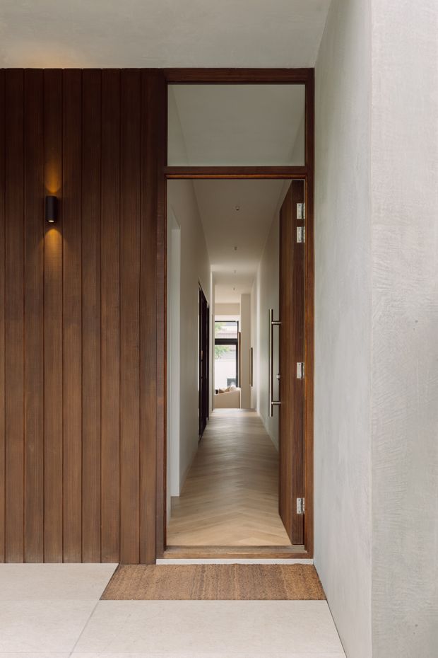 A refined residential entryway viewed from outside, with a tall timber-framed glazed door opening onto a long bright hallway. The entry facade combines vertical dark timber wall panelling with pale textured plaster walls and large light floor tiles. Warm wall lighting, timber door hardware, and a glimpse of seating and glazing at the end of the corridor create a calm, contemporary atmosphere.