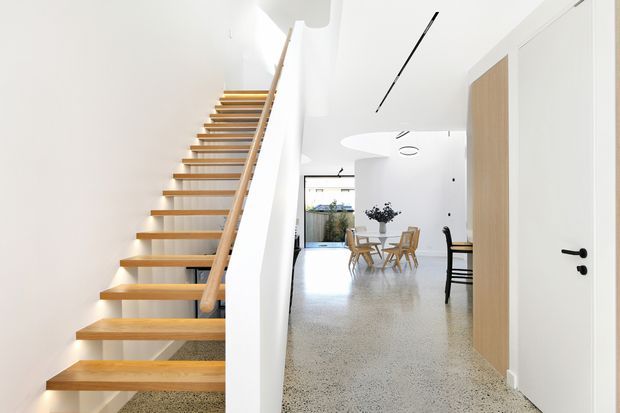 Bright minimalist interior view of a hallway and open-plan dining area, featuring a floating timber staircase with a pale wood handrail, white walls and ceiling, polished terrazzo flooring, concealed linear lighting, and a white door with black hardware. A dining table and chairs are visible beyond the stair and corridor.