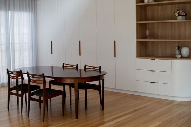 A refined dining area with a dark wood oval dining table and matching mid-century-style chairs, set against full-height white built-in cabinetry. The cabinetry includes warm wood open shelving, white drawers, and slim wood pulls. Timber flooring, sheer curtains, and soft natural light create a warm, understated interior.