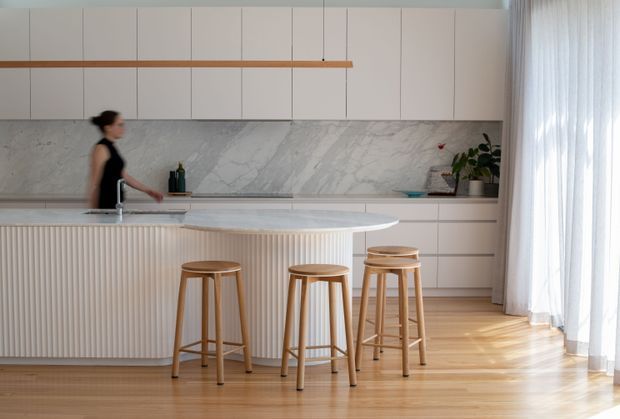 Bright contemporary kitchen with a large white marble-look island featuring a vertically fluted front, three timber bar stools, pale flat-panel cabinetry, a matching marble-look splashback, timber flooring, and sheer full-height curtains. A linear timber pendant hangs above the island.