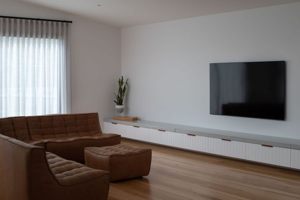 A contemporary living room with a large brown tufted sectional sofa and matching ottoman, a wall-mounted flat-screen television, and a long white built-in media cabinet with a grey countertop. Floor-to-ceiling sheer curtains provide soft daylight, while light timber flooring and white walls create a restrained, uncluttered interior.