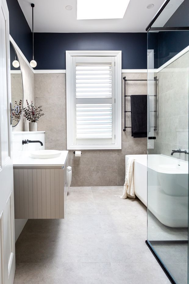 A bright, refined bathroom featuring a freestanding white bathtub behind a frameless glass shower screen, a floating fluted vanity with a white vessel basin, and a black wall-mounted tap. Large pale stone-look tiles cover the floor and lower walls, while the upper walls are painted deep navy with white trim. A shuttered window, black heated towel rail, round mirror, pendant lights, and a skylight complete the space.