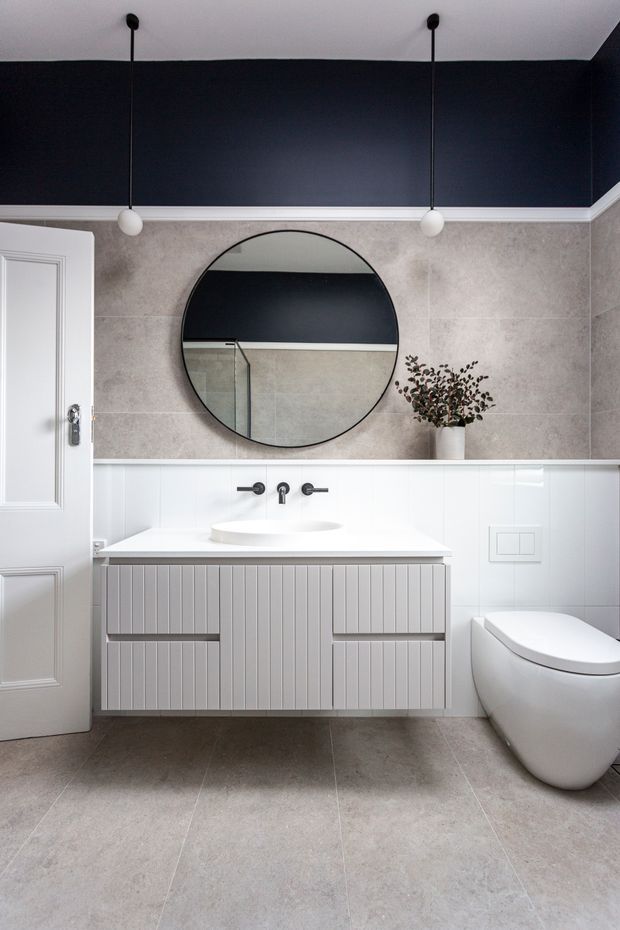A refined contemporary bathroom featuring large grey stone-look floor and wall tiles, white subway-style lower wall tiling, a floating white vertically fluted vanity with integrated basin, matte dark tapware, a wall-hung toilet, and a large round black-framed mirror. The upper wall is painted deep navy with white decorative trim, complemented by two globe pendant lights and a small potted arrangement.