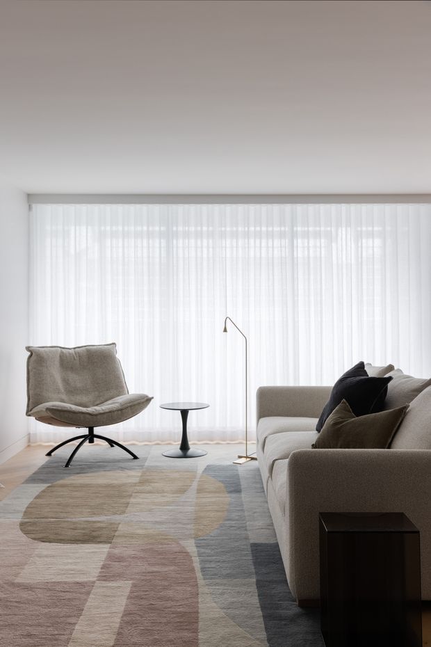 A bright, minimalist living room with a large wall of sheer floor-to-ceiling curtains, a sculptural lounge chair, compact round side table, slim floor lamp, and a neutral sectional sofa. The room features a softly patterned abstract area rug in beige, grey, and muted blush tones, with understated contemporary furnishings and abundant diffused daylight.