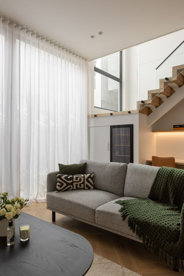 Bright contemporary living room with a large grey sectional sofa, dark rounded coffee table, patterned cushion and textured green throw. Sheer full-height curtains cover expansive glazing, while a sculptural timber stair with glass balustrade rises beside built-in shelving and warm concealed lighting. The room has a clean, refined palette of white, grey, timber and deep green.