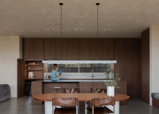 A refined open-plan kitchen and dining area with extensive dark timber cabinetry, a central island with a pale stone benchtop, and a long timber dining table. A broad horizontal window above the sink brings in natural light, while slim linear pendant lights and recessed ceiling lights create a contemporary, understated atmosphere. Built-in shelving and tall storage run along the rear wall, with leather dining chairs and a partial sofa visible.