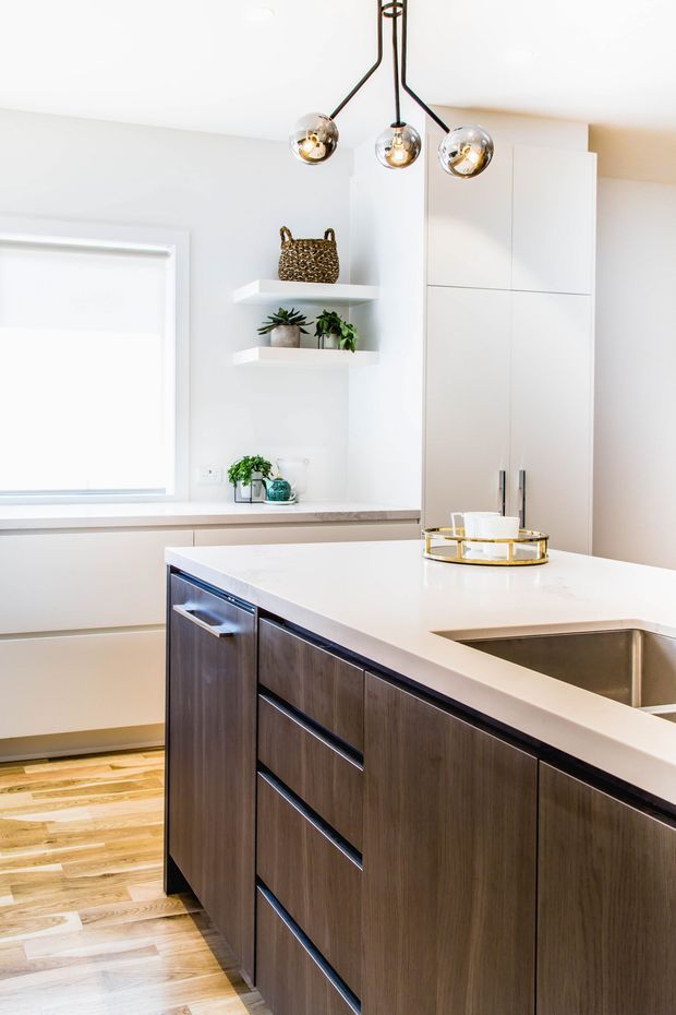 Bright contemporary kitchen featuring a large white waterfall island with an inset stainless-steel sink, dark wood-grain cabinetry, pale engineered-stone benchtops, white full-height storage, open display shelves, light timber flooring, and a sculptural three-globe pendant light.