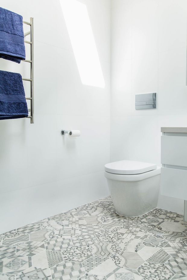 Bright, compact contemporary bathroom featuring white wall tiles, a wall-mounted toilet, floating white vanity cabinetry, a chrome heated towel rail with navy towels, and a decorative patterned grey-and-white tiled floor. The space is illuminated by a strong skylight or clerestory-like opening and has a clean, minimal feel.