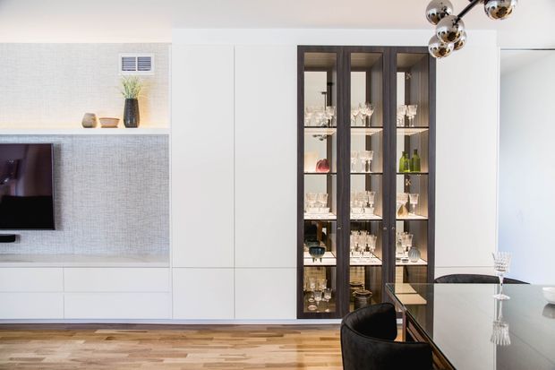Contemporary dining and living area with a built-in white media wall, textured pale wall finish, dark timber-framed glass display cabinetry, and a glossy dining table with dark upholstered chairs. A sculptural chrome spherical pendant hangs above the dining area, while warm timber flooring adds contrast.