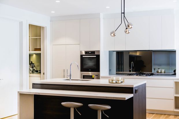 Bright contemporary kitchen featuring a large island with a dark timber-look front, pale stone benchtops, integrated cabinetry, built-in oven, open pantry shelving, and a dark reflective splashback. Three minimalist pendant lights hang above the island, with timber flooring adding warmth.
