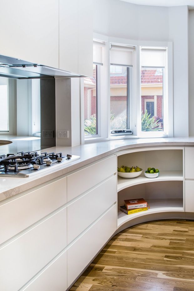 Bright contemporary kitchen featuring white integrated cabinetry, a pale stone benchtop, gas cooktop, mirrored splashback, curved open shelving, and timber flooring. Tall windows with roller blinds provide abundant natural light and views to neighbouring brick buildings.
