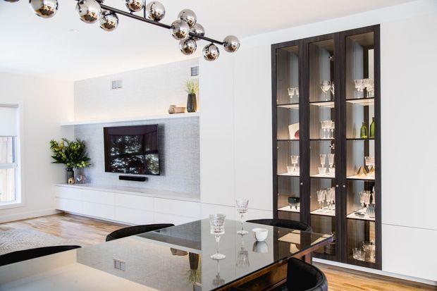 Bright contemporary dining and living area with a glossy dark dining table, black dining chairs, a wall-mounted television set into a textured light feature wall, and a tall dark timber glass-fronted display cabinet. A sculptural multi-globe pendant light hangs overhead, with pale timber flooring and white walls creating a clean, refined interior.