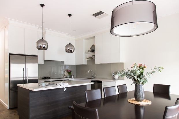 Bright contemporary kitchen and dining area with white flat-panel cabinetry, dark timber-toned island and dining table, stainless-steel appliances, reflective grey splashback, pendant lighting, and a floral arrangement on the table.